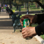 Primer plano de las manos de una persona abriendo una lata de cerveza verde en un parque público con transeúntes difuminados al fondo.