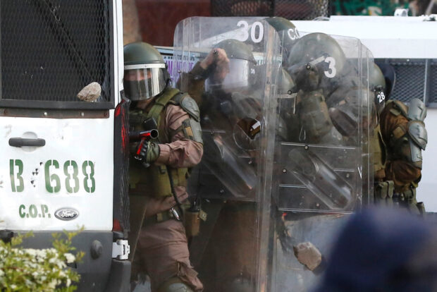 Efectivos de Control de Orden Público (COP) de Carabineros de Chile con cascos y escudos transparentes se resguardan detrás de un vehículo policial blindado durante una manifestación.