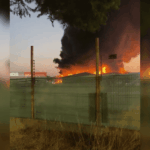 Incendio en un complejo de bodegas industriales con intensas llamas y una densa columna de humo negro elevándose hacia el cielo al atardecer, visto detrás de una reja perimetral.