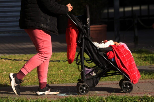 Plano medio de una persona caminando por una vereda mientras empuja un coche de paseo negro con un bebé abrigado con una manta roja