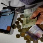 Manos de un adulto mayor sosteniendo billetes de mil y dos mil pesos chilenos sobre un montón de monedas, junto a unos lentes y una calculadora.