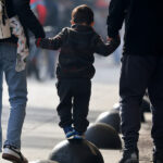 Bono por hijo: Gobierno ingresa con suma urgencia proyecto de ley para el 80% más vulnerable Vista trasera de un niño pequeño tomado de la mano de dos adultos mientras camina y hace equilibrio sobre esferas de concreto en una vereda peatonal urbana.