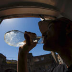 Silueta de perfil de un hombre con gorra bebiendo agua de una botella, con un cielo azul despejado y edificios visibles a través de una ventana al fondo.