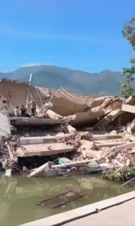 Escombros de un gran edificio de hormigón completamente colapsado junto a un cuerpo de agua, con personas sobre las ruinas.
