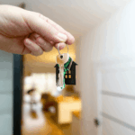 Minvu inicia llamado para el Subsidio de Arriendo (DS 52): ¿cómo postular? Primer plano de una mano sosteniendo un juego de llaves con un llavero negro en forma de casa, con el interior de un departamento o vivienda.