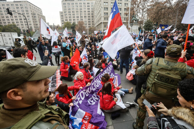 Manifestantes de la salud pública sentados en el suelo sostienen un lienzo, mientras personal de Carabineros está presente en el lugar.