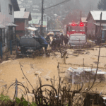 Sistema frontal: Gobierno reporta una noche sin nuevas emergencias y concentra esfuerzos en Corral Calle residencial inundada con agua y barro, se ven vehículos atrapados, ramas caídas y un carro de Bomberos junto a personal de emergencia trabajando bajo la lluvia.