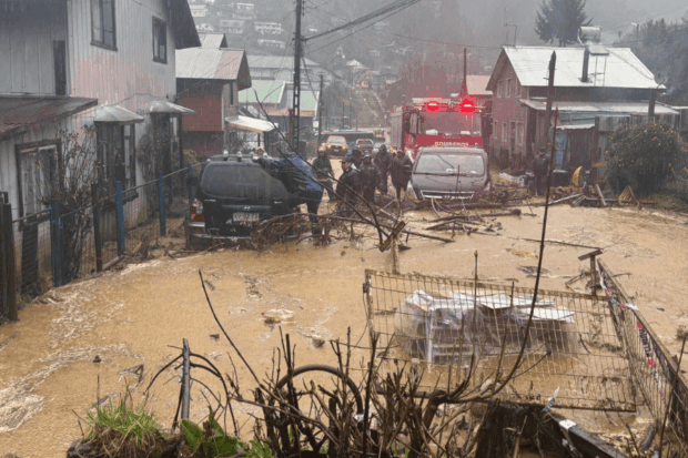 Calle residencial inundada con agua y barro, se ven vehículos atrapados, ramas caídas y un carro de Bomberos junto a personal de emergencia trabajando bajo la lluvia.