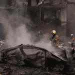 Un ataque ucraniano con más de 400 drones deja 10 heridos en Rusia Vista de los daños causados por un ataque, con bomberos trabajando entre escombros y humo frente a un edificio residencial y un coche calcinado.