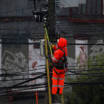 DT recuerda derechos laborales ante corte de suministros: "Sin agua no se puede trabajar" Un trabajador con uniforme de seguridad naranja brillante sube a un poste de electricidad para arreglar cables aéreos durante un día de lluvia.