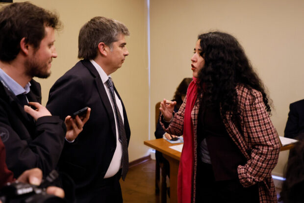 La diputada Daniela Serrano (PC) conversa con dos hombres de traje mientras gesticula con una de sus manos.