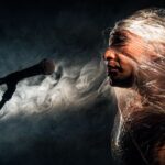 Actor envuelto en plástico se encuentra frente a un micrófono, rodeado de humo y en un escenario oscuro.
