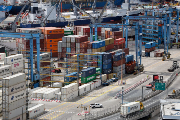 Vista aérea de un terminal portuario con contenedores de carga apilados (Maersk, Hapag-Lloyd), grúas y camiones en operación.
