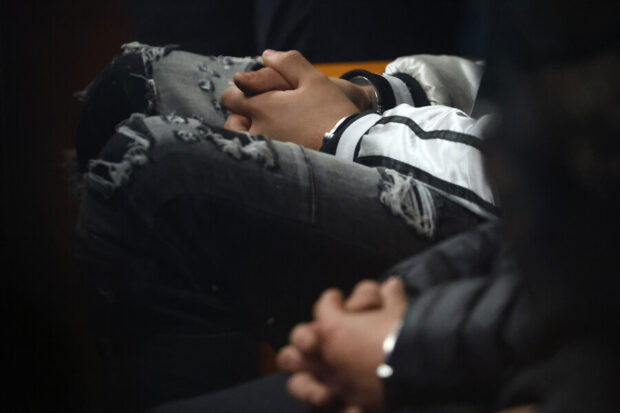 Primer plano de las manos esposadas de dos jovenes durante una audiencia judicial, vistiendo jeans rasgados y chaqueta deportiva.