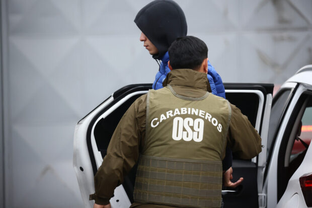 Carabinero escolta a un joven con capucha oscura y chaqueta azul al interior de un vehículo policial blanco.