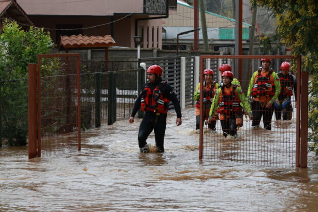 Equipo de rescatistas y personal de emergencia equipados con chalecos salvavidas y cascos rojos caminan con el agua hasta la rodilla por una calle inundada frente a viviendas.