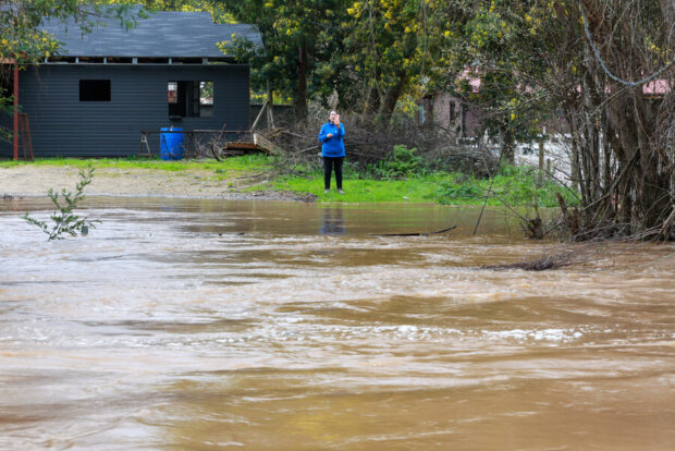 Aumento del caudal de un río desbordado cerca de casas, con una persona observando desde la orilla.