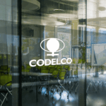 Un despido y amonestaciones: Codelco sanciona a 8 ejecutivos que inflaron registro de producción en 2025 Logo blanco de Codelco sobre un ventanal de vidrio en una oficina moderna con sillas al fondo.