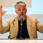 Jorge Baradit: "La Vicaría, la única isla hacia donde nadar en medio del naufragio" Jorge Baradit frente a un micrófono y ambos brazos levantados.
