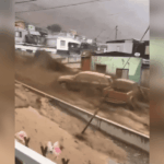 Río de lodo e inundación arrastrando una camioneta frente a casas residenciales en medio de intensas lluvias.