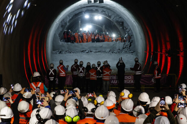 Grupo de trabajadores e ingenieros con cascos blancos y chalecos reflejantes presencian el encuentro de dos frente de excavación en un túnel de metro.