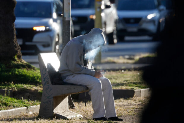 Hombre con capucha gris fuma en una banca al aire libre.