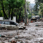 Gobierno defiende rápida respuesta por aluvión en Las Condes tras críticas por diferencias con emergencias anteriores Vehículos dañados y semi enterrados en lodo, ramas y piedras sobre una calle tras el paso de un aluvión.