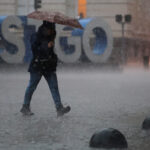 Por lluvias y posibles tormentas eléctricas: Senapred declara alerta temprana preventiva para la RM Hombre con paragua camina en una calle urbana, donde llueve y cae granito. Detrás se ven las letras STGO.